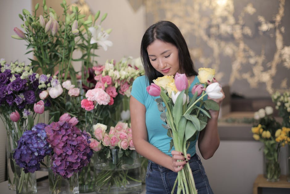 A young woman with shoulder-length dark hair and light skin is examining a bouquet of fresh tulips in a flower shop. She is wearing a light blue top and jeans, holding a mixed arrangement of pink, whi