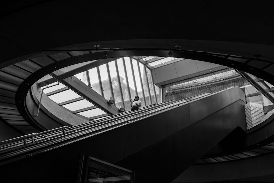 A black and white photograph of an interior stairwell with a spiral design, featuring modern architectural elements including large glass panels allowing natural light to fill the space. A woman is st