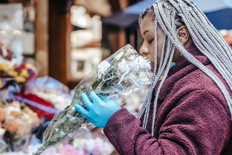 A woman with braided silver hair, dressed in a maroon fleece jacket and wearing a blue glove, holds a bouquet of fresh white roses wrapped in clear plastic at a flower stall. The arrangement features