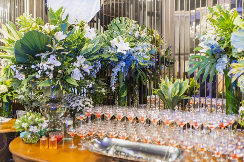 A large floral arrangement featuring white lilies, white roses, and greenery, including broad-leafed tropical plants and monstera leaves, is displayed on a table with a black metal railing in the back