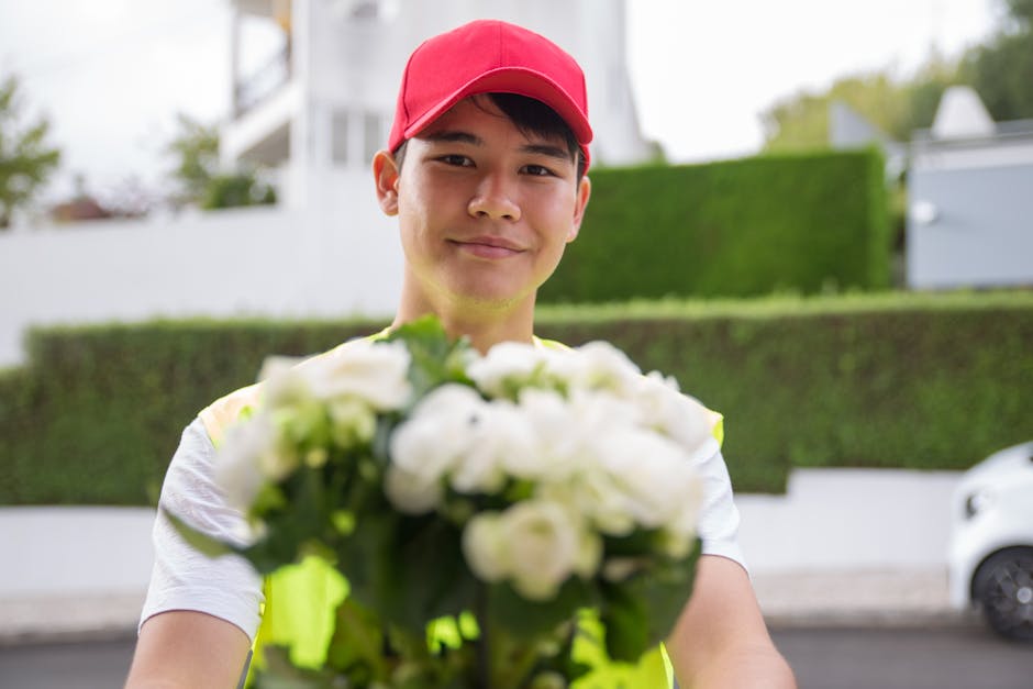 A delivery person with a friendly expression, wearing a red cap and high-visibility vest, holds out a bouquet of white roses, hydrangeas, and green foliage in front of him. The bouquet is arranged in 