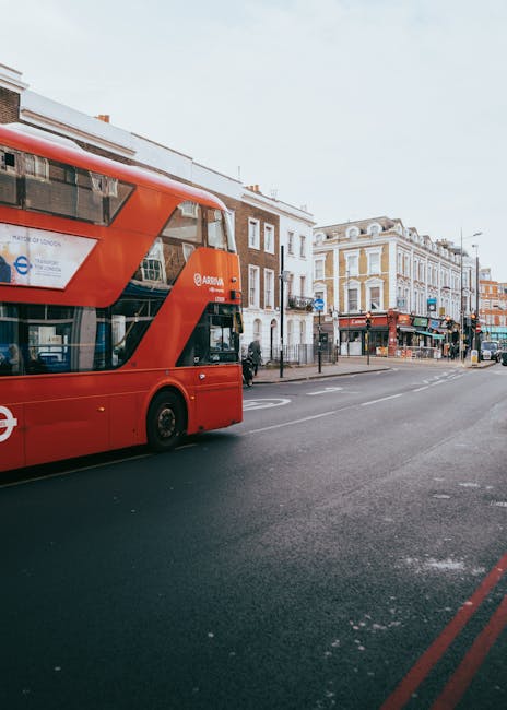 A double-decker red London bus traveling along a busy street in Westminster, with traditional Georgian-style buildings lining the sidewalk. The scene captures a typical city setting during daytime, wi