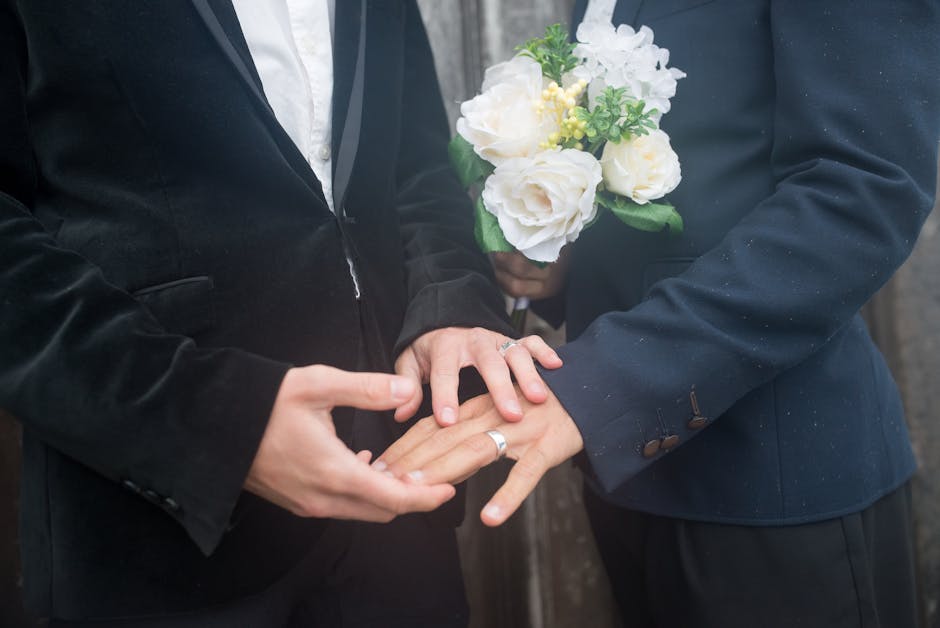 A person in a dark suit and white shirt holds a bouquet of fresh flowers featuring white roses, hydrangeas, and yellow accents with green foliage, wrapped in clear cellophane. Another individual, also