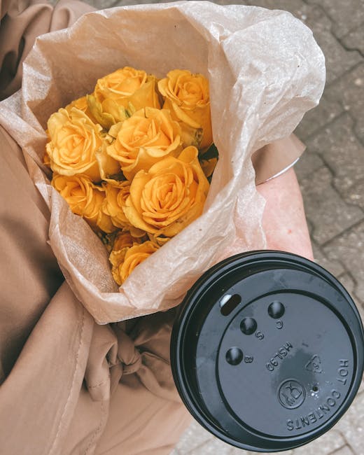 A bouquet of fresh, vibrant yellow roses with tightly packed petals and green stems is wrapped in light brown kraft paper and white tissue paper. The bouquet is held by a person wearing a beige coat o
