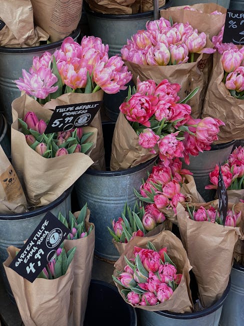 A display of fresh pink tulips in full bloom, arranged in brown paper wraps and placed inside metal buckets at a flower market. The tulips have vibrant petals with varying shades of pink, some more pa