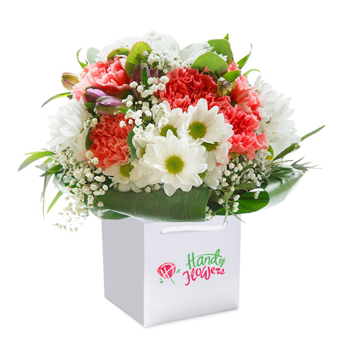 Resting upright in a clean, minimalist white square gift bag, a lavishly crafted bouquet reveals itself in vivid detail-a hallmark of Mayfair's finest floristry. The focal point is a graceful interplay of coral and soft pink carnations, their exuberantly ruffled petals forming plush layers of warmth and romance. Bright white daisies, each with a vivid yellow-green heart, dot the arrangement, infusing the display with a cheerful, optimistic energy reflective of spring blossoms found around Grosvenor Square. Delicate sprigs of snowy gypsophila are woven throughout, their fine, cloudlike clusters introducing lightness and a sense of celebration. Tucked discreetly to the left, small buds show hints of purple blush, waiting to bloom and adding a note of intrigue and depth to the arrangement's shape. Deep green, glossy foliage wraps generously around the bouquet's base, its broad leaves creating a natural border that anchors and elevates the blooms above. All elements are arranged to achieve a rounded, abundant silhouette that speaks of freshness and skilled artistry. The plain white bag, finished with elegant ribbon handles, is designed for effortless carrying-perhaps en route to a birthday dinner by Bond Street or delivered to a loved one's address nearby. With the entire scene set against a stark white background, the flowers' vitality and colour radiate; this bouquet perfectly embodies the luxury and refinement for which Mayfair is known, making it suitable for birthdays, anniversaries, or any moment deserving of a distinguished floral gift.