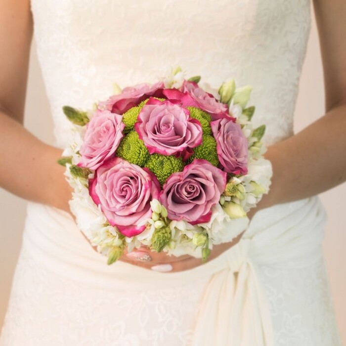 A close, intimate photograph reveals a bride from Mayfair gently clutching her artfully arranged bouquet against the backdrop of her elegant, vintage lace wedding gown-its delicately patterned overlays echoing the timeless charm of Berkeley Square's classic architecture. The bouquet commands attention at the centre: fat, fully opened roses in shades of dusty rose and mauve, their petals feathered with hints of fuchsia, evoke both softness and visual intrigue. Nestled between the central roses, a ring of vivid green button chrysanthemums introduces a bright, lively energy, their round, sculpted heads and zesty colour evoking the fresh, vibrant gardens dotted along Curzon Street. Encircling all are clusters of diminutive, snow-white blossoms-possibly freesia or petite spray roses-with their green buds delicately scattered among them, contributing subtle scent and a sense of fresh beginning. The bride's hands are visible just below the bouquet, her nails neatly manicured in pale pink, with a subtle, sparkling accent adding an elegant touch that matches the overall sophistication of the look. Soft, even lighting brings out the layers of lace and the variegated gloss of each petal, making every texture and hue stand out. The composition is one of harmony: pastel roses, crisp white flowers, and vivid green mums unite in a joyous, romantic bundle, the perfect highlight for wedding celebrations in the heart of Mayfair. The effect is at once classic and contemporary-an impeccable choice for articles or guides dedicated to distinguished wedding florals, palette ideas, and bridal styling for central London events.