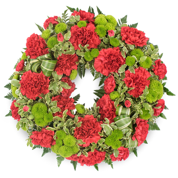 A generously proportioned funeral wreath, perfectly circular and richly textured, is pictured against a bright white background that highlights the interplay of red, green, and soft shadow. The ring is densely packed with flowers, giving it a substantial, almost sculptural presence, as if no space is left untouched by petals or foliage. Dominating the design are deep red carnations, their many-layered, frilled petals creating a velvety surface that feels both classic and deeply comforting. The carnations are arranged evenly around the wreath, yet they vary slightly in size and openness, so the composition feels organic rather than mechanical. Between these crimson blooms, clusters of lime green button chrysanthemums, or pompon mums, appear like luminous beads of colour, each small flower forming a perfectly rounded dome of tiny petals. The chartreuse hue offers a vivid contrast, bringing a subtle sense of hope and renewal to the tribute. Underpinning the flowers is an abundant framework of greenery: darker fern fronds, probably leatherleaf, radiate outwards with feathery detail, while variegated pittosporum leaves with creamy edges weave in and out, adding lighter notes that prevent the wreath from feeling too heavy. Discreet loops of a striped green ribbon-pale green crossed with deeper green diagonals-are tucked into the foliage, particularly visible along one side, adding an elegant flourish that remains in harmony with the natural elements. The flawless circle of the wreath symbolises continuity, remembrance, and the unbroken bond of love. Crafted with quiet care in Mayfair, close to the dignified avenues around Berkeley Square, this arrangement feels ideal for a funeral or memorial service where family and friends wish to offer a refined, heartfelt expression of sympathy.