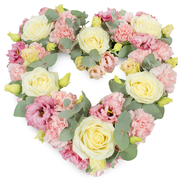 In this image, a generous heart-shaped wreath is presented against a clean, white background, its pastel flowers forming a complete, symmetrical heart that seems to float in quiet stillness, like a moment of reflection in Mayfair's side streets. The heart is densely packed, every space filled with petals and foliage so that the design appears rich yet softly blended. Dominant within the arrangement are large roses in shades of cream and pale butter yellow, their velvety petals curled tightly at the centre and unfurling toward the edges, giving each bloom a sense of poise and dignity. Threaded between these roses are many clusters of light pink carnations, their frilled, delicate petals layered densely, adding a soft, billowing texture that gently breaks up the roundness of the roses. Deeper pinks and mauve tones appear in the lisianthus flowers, whose more open, gently fluted petals and darker centres introduce a quiet depth and hint of evening shadow. Small yellowish-green buds peep from within the heart, suggesting freshness and the tender beginning of new blooms. The entire tribute is framed and intertwined with silvery-green eucalyptus foliage, its smooth, rounded leaves adding subtle movement and a cool, calming tone. The palette of blush pink, cream, and muted green evokes tenderness, sympathy, and a deep, steady love. This elegant heart wreath would be at home at a Mayfair funeral or memorial, perhaps resting by the entrance of a church near Hanover Square or delivered as a condolence tribute to a townhouse just off Park Lane, conveying care and remembrance through thoughtful floristry.