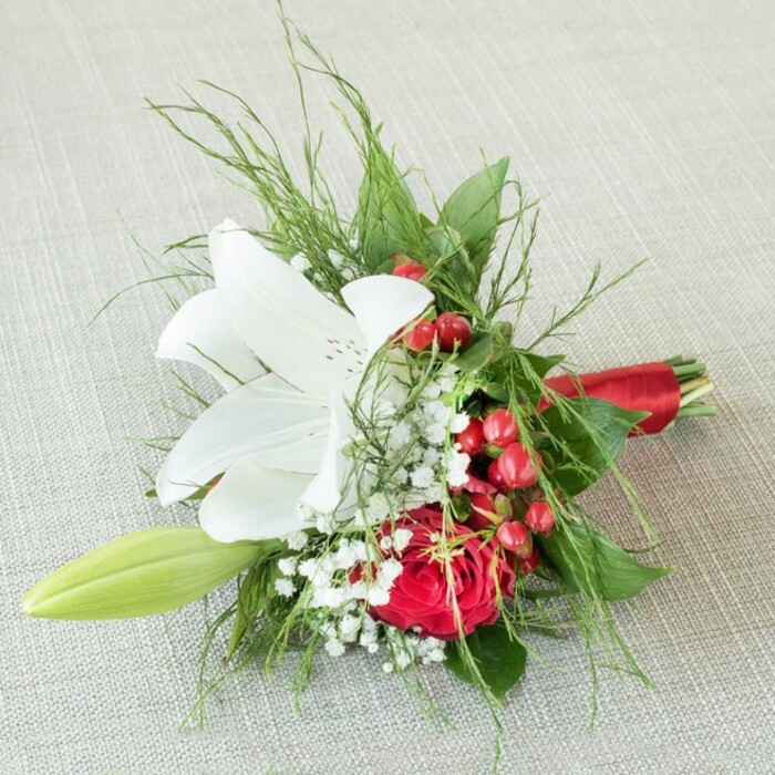 A beautifully composed bouquet, likely for a bridesmaid or cherished guest, is displayed from above on an understated, textured beige fabric that echoes Mayfair's refined aesthetic. Dominating the arrangement is a luminous white lily-its petals fully unfurled and dusted with tawny red freckles, drawing focus to the blossom's yellow-gold centre. Nestled nearby, a deeply saturated red rose with velvety petals adds drama and tenderness, embodying affection and the grandeur of Mayfair's renowned floristry. White sprigs of gypsophila dot the bouquet, imparting delicacy and a breath of the English countryside, while clusters of vibrant red berries bring joyful colour reminiscent of local Mayfair festivities and garden parties. A pale green lily bud arches beside the main blooms, symbolising new beginnings and the promise of growth. Fine feathery fern leaves and wide, dark green foliage frame the composition, enhancing its depth and lushness. The stems are presented neatly, bound by a silky scarlet ribbon-a nod to the bouquet's festive mood. This expertly arranged piece radiates emotional warmth and celebration, making it a fitting accent for weddings, anniversaries, or elegant gatherings in Mayfair, each flower chosen for its symbolism and freshness, ready for prompt delivery to addresses across central London.