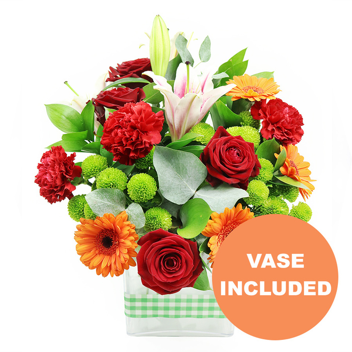 A vibrant mixed bouquet brimming with saturated reds, oranges, whites, and greens is captured here in crisp detail, as though freshly arranged in a Mayfair studio. The composition is densely packed, giving an impression of abundance and celebration. Deep crimson roses form one of the key focal points; their petals are still tightly layered, creating exquisite spirals of velvety red that hint at long-lasting freshness. Around them, ruffled red and red-orange carnations provide a softer, more feathery texture, their gently frayed edges catching the light and adding a sense of movement. Striking orange gerbera daisies punctuate the design, each one a bold disc of colour with a dark central eye, like miniature suns radiating warmth and cheer. Rising above this main cluster are graceful white Oriental lilies; some are fully open, their trumpet-shaped blooms displaying pale pink stripes along the interior of the petals and golden stamens dusted with pollen, while others are closed, sculptural buds suggesting blooms yet to unfold. Nestled between the larger flowers, button-like lime-green pompom chrysanthemums add crisp, rounded shapes and a zesty shot of colour, balancing the intense warm tones. The lush greenery consists of broad, glossy dark green leaves and soft, silvery eucalyptus sprays, which create depth, contrast, and a gentle, almost woodland feel reminiscent of strolling through nearby Hyde Park. All of this is presented in a sleek, clear rectangular glass vase that reveals the tangle of stems within, its base wrapped with a light green and white gingham ribbon for a subtly rustic accent. In the lower right corner of the picture, a bright orange circular badge with VASE INCLUDED in clear white capital letters overlaps the edge, signalling that the stylish vase forms part of this ready-made gift, ideal for birthdays, thank-you gestures, or congratulations delivered across Mayfair.