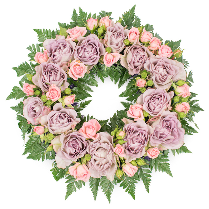 Set against a clean, white backdrop, a meticulously designed funeral wreath is captured from directly above, allowing its full circular symmetry to be seen in one tranquil view. The ring is richly composed of large roses in muted lavender, dusty mauve, and soft greyish-purple tones, each bloom fully opened to reveal tightly layered petals that spiral gracefully into a shaded centre. These elegant roses are carefully spaced so their rounded forms create a gentle rhythm around the wreath, never crowded yet seamlessly connected. Between them, clusters of smaller pastel pink spray roses and partially opened rosebuds are interwoven, their lighter colour infusing the arrangement with a tender warmth that softens the more subdued purples. Beneath and around the flowers, a dense bed of dark green fern fronds forms a continuous, feathery base, the serrated leaves splaying out in all directions to create a lush, living frame. Occasional glimpses of deeper, shadowy greenery and tiny dark buds tucked among the petals give the design extra depth, as if the wreath were drawn from a peaceful woodland garden. The lighting is soft and even, with no harsh contrasts, highlighting the natural textures and colour gradations across every bloom. This is the sort of refined tribute that might be chosen for a Mayfair farewell near Shepherd Market or a quiet service by Green Park, where families value understated, classical beauty. The craftsmanship suggests an experienced florist working with calm concentration, building a floral circle that conveys sympathy, gratitude, and enduring remembrance, ideal for funerals, memorials, or dignified commemorations of a cherished life.