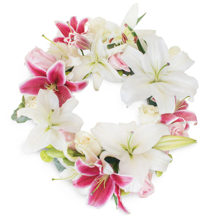 An elegant and meaningful floral wreath takes center stage, exuding the refined artistry that characterizes so many of Mayfair's beloved traditions. This circular tribute features lush, star-shaped white Oriental lilies as its primary focus-each bloom pristine and luminous, their petals arching gracefully around prominent pistils and soft yellow stamens that catch hints of daylight. Weaved seamlessly among them, bold fuchsia and magenta-pink Stargazer lilies create dynamic accents, their intense color and gentle speckling offering dramatic counterpoints within the design. Spheres of pale blush and creamy white roses, some tightly furled and others unfurling into delicate softness, nestle between the lilies, softening the arrangement with romantic texture and muted pastel hues. Discreet sprigs of fresh, green foliage-perhaps young eucalyptus or the smooth leaves of the lilies themselves-peek through from behind, contributing subtle depth and a whisper of woodland fragrance. Every element in the ring is curated with thoughtful intention, forming a continuous, unbroken circle that eloquently communicates unity, eternity, and the cycle of remembrance. Set against a striking white background, the wreath appears luminous and dignified, a reflection of the timeless beauty found in Mayfair's historic streetscapes and tranquil hidden gardens. Appropriate for honorific services or private gatherings near Grosvenor Square, the overall effect is one of gentle celebration and deep commemoration-a floral creation that brings grace, solace, and connection to local families in their times of memory and tribute.