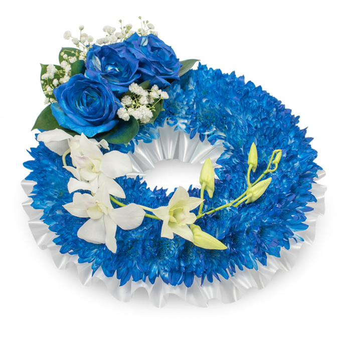 Presented against a simple white background, this funeral wreath is a carefully balanced composition of deep blue and pure white tones, conceived for dignified remembrance in the heart of Mayfair. The wreath's core is densely packed with countless small royal-blue chrysanthemums, their fine, radiating petals building a thick, cushiony ring that feels substantial and consoling to the eye. A pleated white satin ribbon lines the inner opening, its soft folds and subtle luster creating a dignified frame that offsets the intensity of the blue blooms. Near the upper-left curve, a trio of large blue roses opens fully, their silky, layered petals a richer sapphire that draws the gaze; tiny sprays of white gypsophila and touches of dark green foliage sit nearby, adding air and visual relief. Flowing across the lower portion of the wreath is a cascade of pristine white orchids - likely Phalaenopsis with their classic rounded petals - arranged to arch gently from the mid-left toward the lower right, interspersed with pale green orchid buds hinting at new life. The contrast between the orchids' smooth, luminous petals and the chrysanthemums' textured surfaces creates a quiet, tactile harmony. In soft, cool daylight the piece reads as composed and respectful, its pale orchid scent faint and clean against the earthier mum notes. As a Mayfair tribute suitable for services around Grosvenor Square, Berkeley Square or private rooms near Bond Street, this wreath was carefully prepared with the attention to detail that clients expect from Mayfair Florist, conveying sympathy, purity, and enduring remembrance.